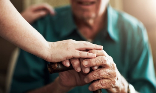 Elderly man  sitting down and holding his cane while a young person holds his hand