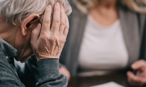 An elderly man looking troubled with his right hand placed on his head.