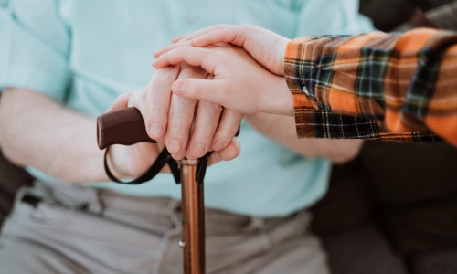 Elderly man  sitting down and holding his cane with two younger hands on top of his
