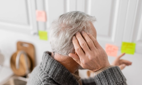 A grey-haired senior man touches his head and points to a blurred sticky note in the foreground.