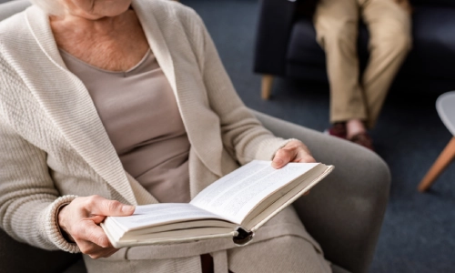 An elderly woman sitting comfortably on a sofa chair while reading a book.