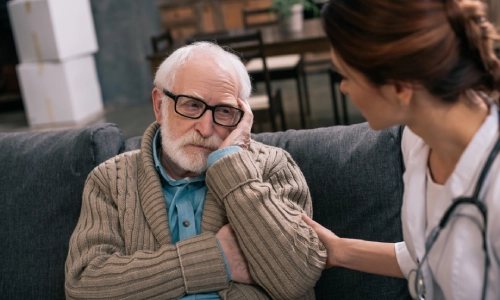 A compassionate healthcare professional is listening attentively to an elderly man sitting on a sofa who appears to be lost in thought.