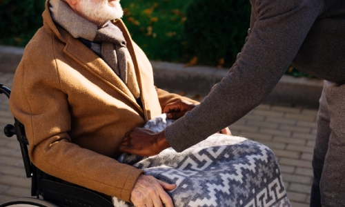 A young man gently tukcs a warm blanket around the knee of an elderly man in a wheelchair.