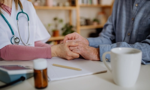 A female doctor holding the hand of a senior man while discussing his medical condition over a cup of coffee and a bottle of medicine on the table.