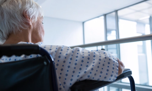 An elderly woman in a hospital gown is sitting in a wheelchair while gazing out of a large window.