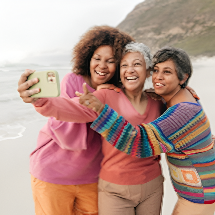 Three women smiling and taking a selfie together on a beach with ocean and cliffs in the background.
