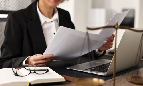 A New Jersey Medicaid planning attorney holding files in both hands while sitting in front of an open laptop with her glasses on the side of the table